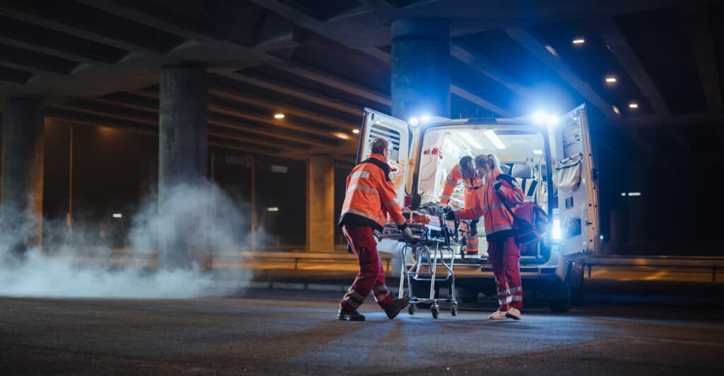 Paramédics chargent un patient sur une civière dans une ambulance la nuit sous un pont avec de la fumée.