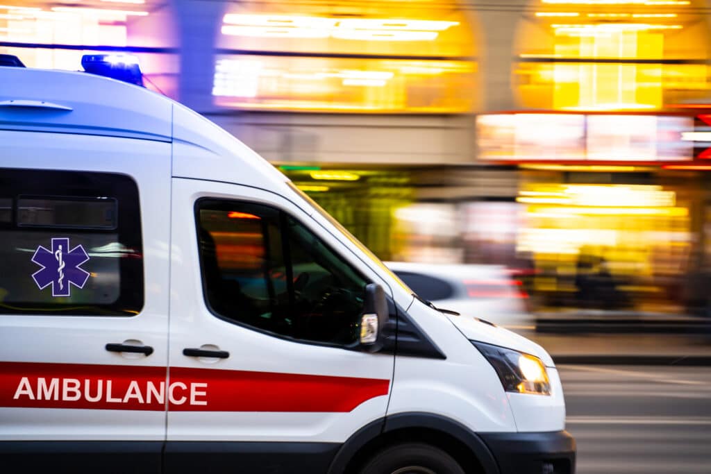 Ambulance blanche avec sirène bleue clignotante et texte rouge "AMBULANCE", filant en urgence dans la ville.