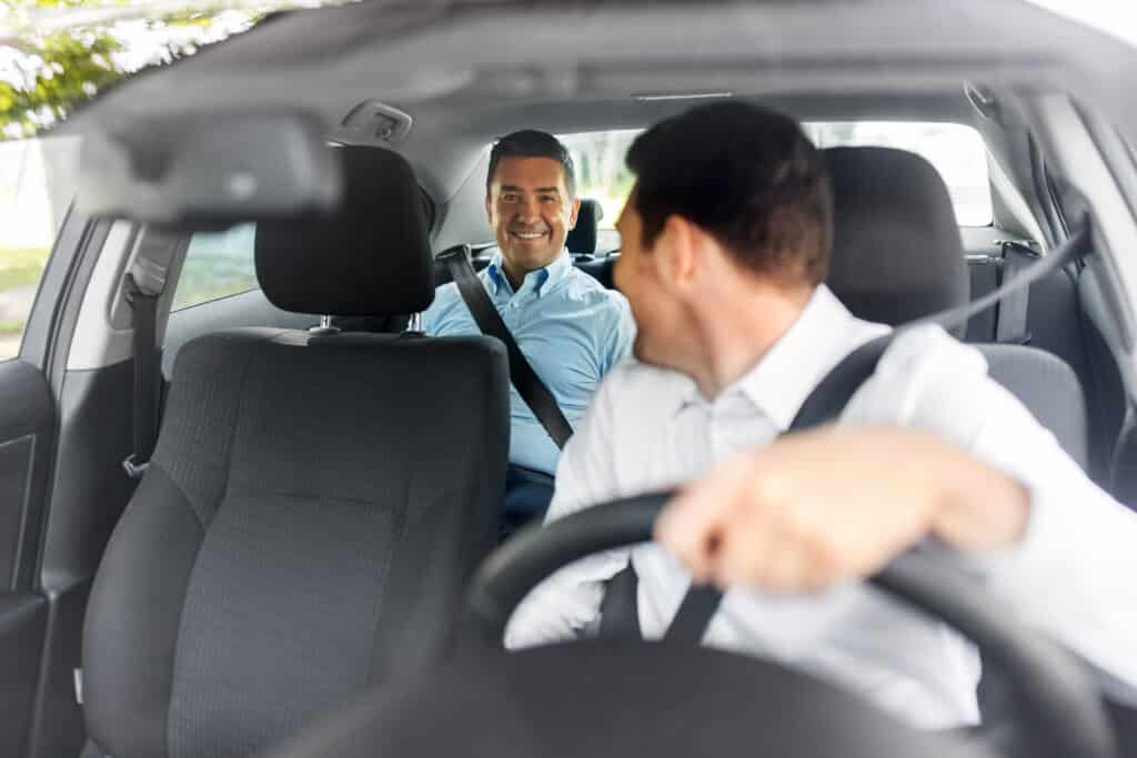 Chauffeur de voiture se tournant vers un passager souriant à l'arrière. Les deux hommes portent leur ceinture de sécurité dans l'habitacle.